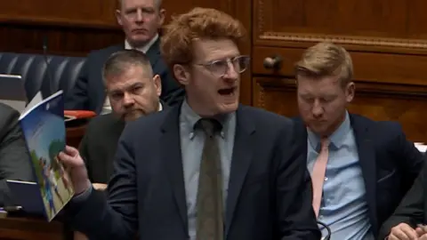 Matthew O'Toole holding the programme for government paper as he speaks to the assembly in Stormont. He has ginger hair, wears clear glasses, and is wearing a dark coloured suit. 