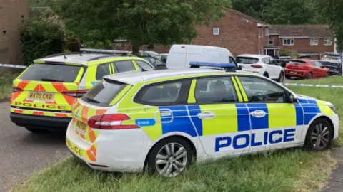Two parked police vehicles in front of police tape cordoning off an area. There are houses and more cars parked behind the police tape. 