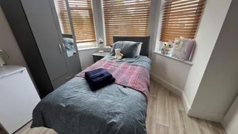 Small room with single bed infront of window with wooden blinds and grey wardrobe at the side