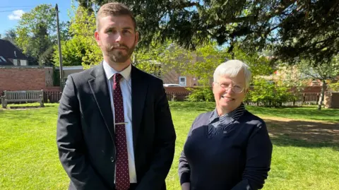 BBC Two people on a sunny day in a park, the man wears a dark suit and burgundy tie, the woman wears a navy jumper and has grey hair and glasses