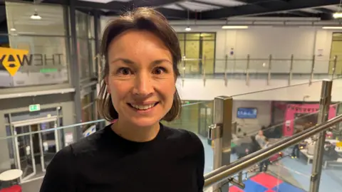 A middle aged woman smiling a the camera. She has short hair and is standing on a balcony. She is wearing a black t-shirt. Down below we can see young people taking part in activities. 