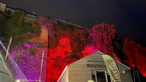 The ivy-covered wall of Newquay Harbour is lit up with red, purple and pink lighting. In the foreground is a building with the letters BOATHOUSE on it and a flag advertising Cornish Rattler.