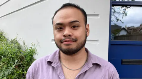 BBC News Japeth Monzon, wearing an open necked lilac shirt, a a silver necklace and a hoop in his nose piercing. He has short dark hair and facial hair and is smiling at the camera. Behind him, is a blue door to a house and a white wall with greenery up to it.
