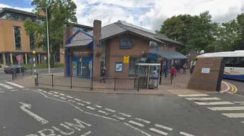 A screengrab of a Google Maps image of Mountjoy Road in Omagh, outside the bus depot.  The two-storey building is constructed of brown brick and it has a roof which slopes at various angles.  There is a bus parked in the car park outside and there is a tall, yellow-brick building in the background. 