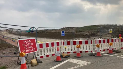 Laura Coffey/BBC Plastic cones and fencing run across a road. There is a sign that says road closed to all vehicles. Behind that is a high metal fence with warning signs on it. In the background a digger can be seen.