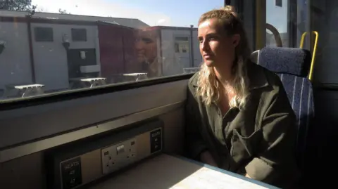 A woman sitting inside a train, looking out of the window as it goes past some industrial units, with her reflection in the glass.