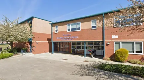 Google A brown and yellow brick two-storey building with bushes and a large cherry blossom tree. A sign above the entrance says Welcome to Hilbre High School