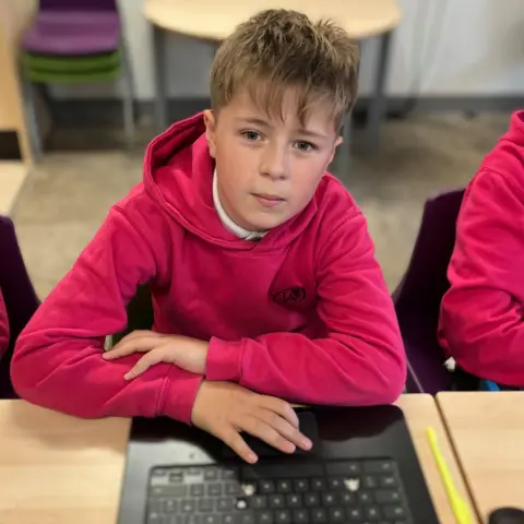 Samuel sitting at a classroom desk with a laptop. He is wearing a deep pink hoodie.