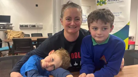 Alun (left) with his mum and brother. They all look at the camera. They are sat in a room at a table. Alun arms are across the table and his head is lying on top of his arms. He has a blue top on, blue eyes and blonde hair. Eleri has a black top and dark brown hair. On the right is Alun's brother who also has a blue top on, it is a school jumper and he has dark brown, slightly wavy hair.