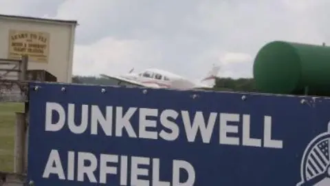 A blue and white Dunkeswell Airfield sign. A small plane is in the background.