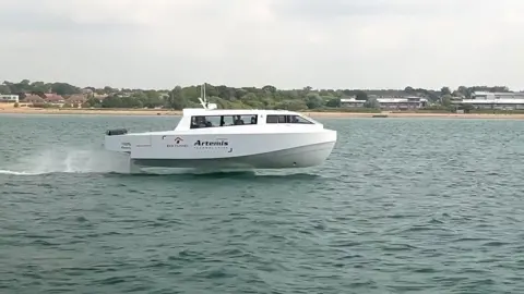 A small, sleek white vessel traveling across the water. Its hull is lifted out of the water and it appears to be gliding. In the background is the shore, with sand giving way to trees and buildings