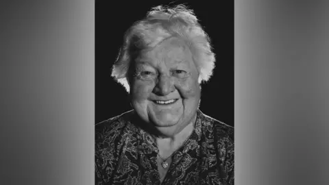 Colin Potsig A black and white photo of a woman with white, short hair smiling into the camera in front of a black background. She is smiling with her teeth and has a heart shaped necklace on with a patterned shirt.
