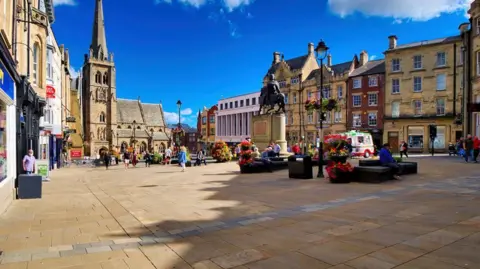 Geograph/David Dixon Durham Market Place on a sunny day half in shade. There are not many people around and an ice cream van can be seen.
