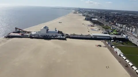Jamie Niblock/BBC A drone image taken from sideways on to the pier. It extends for metres across the beach, with a white theatre building and a slide towards the end of the pier. A stretch of green gardens and white shelters border the promenade.