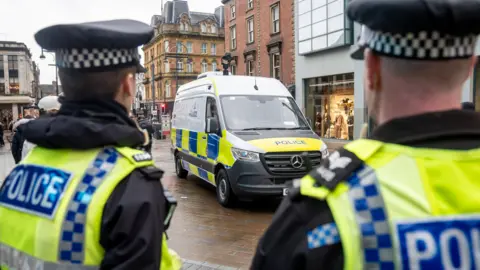 Two police officers stand with their backs to the camera, looking down Briggate in Leeds. They are wearing high-vis vests. They are looking at a police van, which is parked next to a shop.