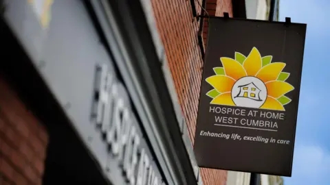 Hospice at Home West Cumbria Close-up image of a sign with the charity's name and logo hanging on a wall. The sign, which has a black background, reads Hospice at Home West Cumbria, enhancing life, excelling in care. The logo has a house at its centre in a white circle, and is surrounded by yellow and green petals.