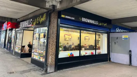 The outside of a cafe in Brownhills. It faces on to a paved area under the canopy of a 60s-style shopping mall.