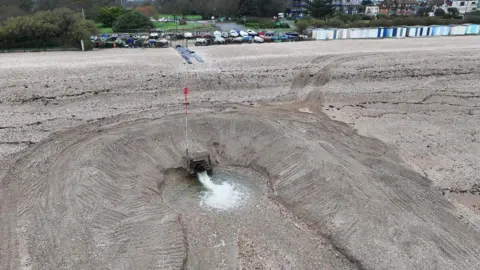 Eddie Mitchell An aerial photo of an outfall pipe spewing out water with shingle around it about 100 metres away from Fish Lane which had flooding. The pipe is surrounded by a mound of shine and the water can be seen draining in two channels out to sea. Cars can be seen parked on the seafront and there are beach huts