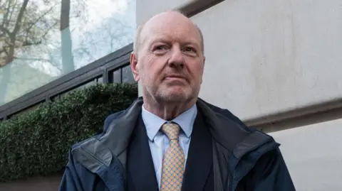 Sir Alan Bates standing outside of the official inquiry into the Horizon IT scandal, wearing a navy rain mac coat over a navy suit and blue dress shirt, accompanied by an orange tie with a green blue and yellow circled pattern