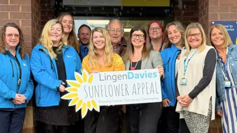 Nine women and three men, some of them wearing blue jackets, holding a large sign which reads Lingen Davies Sunflower appeal