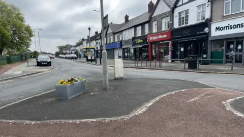 Picture of Childwall Abbey Road which is due to close as part of these plans. The picture is taken from a traffic island, almost a roundabout, whilst no cars are seen on the road. Shops line the street to the right.