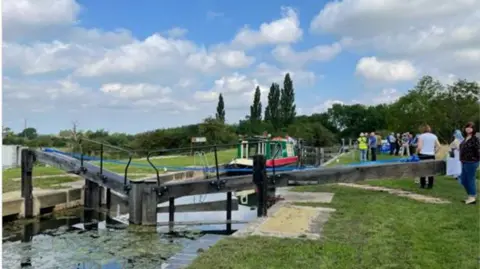 A lock gate of a canal with grassy banks either side and a barge on the waterway, taken on a sunny day with blue skies.