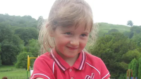 PA Media April Jones, aged five, who has light brown hair pulled back in a ponytail. She has a whispy full fringe pushed to the side. She has brown eyes and smiles at the camera. Behind her a large green wood can be seen. She seems to be in a playground as the corner of a slide can be seen over her shoulder