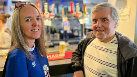 Emma O'Shaughnessy, with long straight blonde hair and wearing an Everton top, and her father Kevin, with a grey jacket and white t-shirt, smile at the camera with a bar in the backdrop