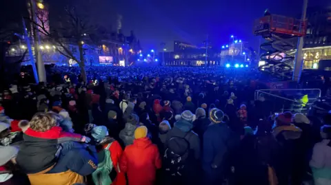 Crowds in Bradford City Park, wearing coats and hats.