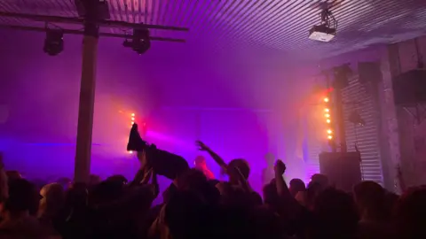 Lucy Blackledge Inside a club. The lights are purple and it is busy. A man can be seen being carried by the crowd.