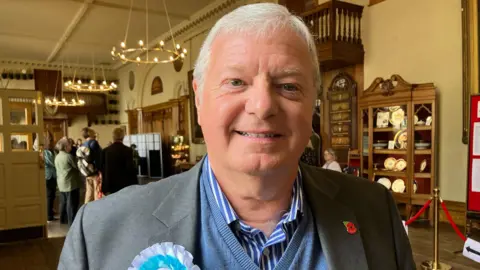A man with short white hair smiles at the camera. He is wearing a blue and white striped shirt under a blue V-neck sweatshirt and a grey suit jacket. A poppy badge is on his left lapel and a blue and white rosette is on the other.