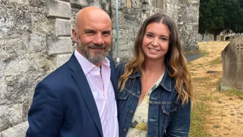 Hannah Gray/BBC A man in a blue suit and a pink shirt. He is standing with a woman in a denim jacket and yellow shirt. They are standing next to a church.
