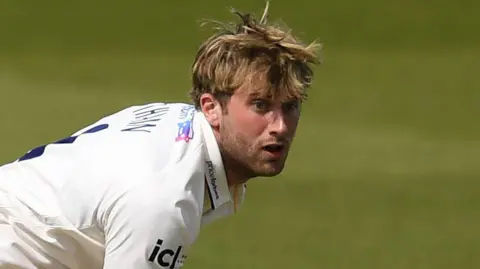 Essex cricketer Noah Thain, who is in a white Essex Cricket jersey, reacts as he bowls during a County Championship match.