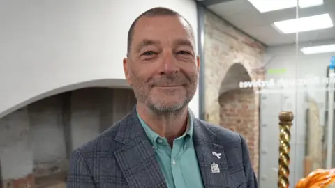 Qays Najm/BBC Simon Ring, a man who is standing up looking directly at the camera and smiling. He is wearing a mint green shirt with a grey blazer over the top.