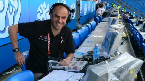 Tony Jones Tony Jones wearing a black polo shirt and headset while sitting in the press box at a Euro 2008 match. He is smiling and surrounded by blue seats, with TV monitors and notepads in front of him.