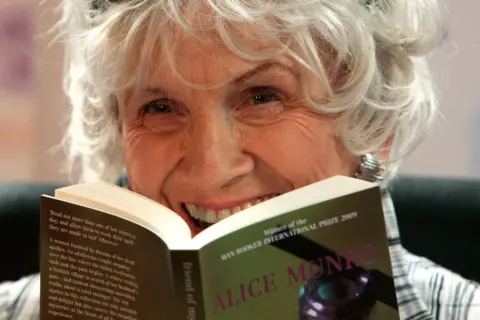 Getty Images Alice Munro is photographed close up and laughing, holding one of her own paperbacks open in front of her. She has short grey slightly wild hair. 