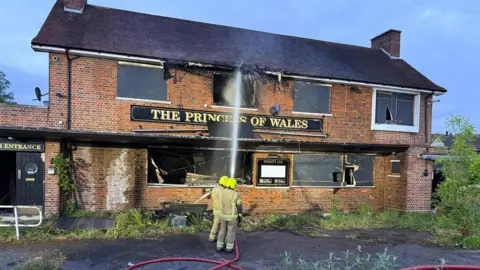 Essex County Fire and Rescue Service Two firefighters with their backs to the camera spraying water from a hose at a derelict pub called the Princess of Wales. 
