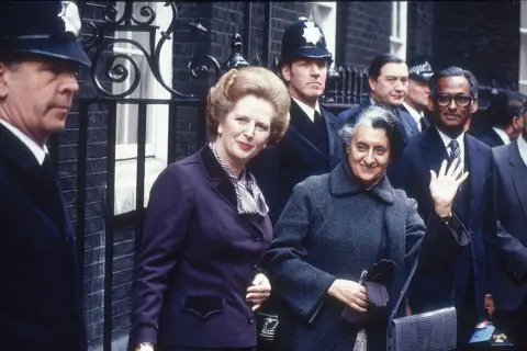 Raghu Rai/The India Today Group via Getty Images A photograph by Raghu Rai that shows Indira Gandhi visits Prime Minister Margaret Thatcher in London, England. Both of them are wearing warm coats and can be seen surrounded by security personnel.