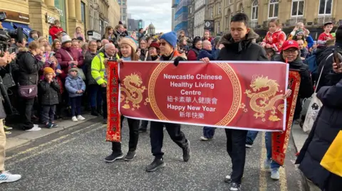 Lots of people in Newcastle City Centre are watching the start of a parade which is led by a banner saying Newcastle Chinese Health Living Centre. Behind the streets are full of people.