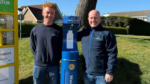 Jersey Water Two men - Sam Allix and Martin Skyba - from Jersey Water wearing Jersey Water clothing stand either side of a blue water refill station installed in the St John's Village area of Jersey. The station is in front of a grass bank and a tree.