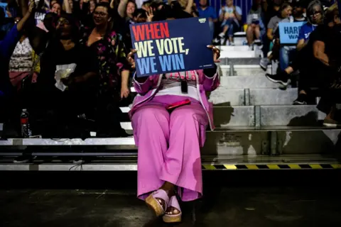 Emily Elconin/REUTERS An woman in a pink suit, sitting in a crowd, holds a sign which reads WHEN WE VOTE, WE WIN