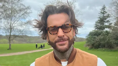 Pete Wickes has a big floppy brown fringe which is windswept, thick framed glasses and is wearing a brown vest jacket. Behind him is the grass lawns of the country park and trees.