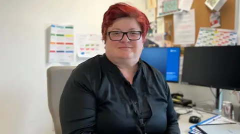 BBC Dr Elisabet Gomes Dos Santos is sitting at her desk and is smiling at the camera. She has short red hair and is wearing a pair of glasses and a black shirt with a lanyard around her neck. There are two monitors on her desk and a few plastic files. On the wall in front of the desk is a noticeboard with various papers pinned to it and there are some pinned directly on the wall to her left.