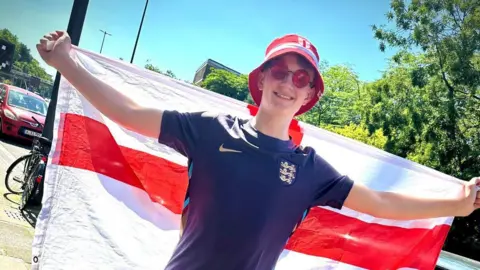 Family photo A boy in a red hat, red sunglasses and dark blue football shirt, holding a large cross of St George flag on his back
