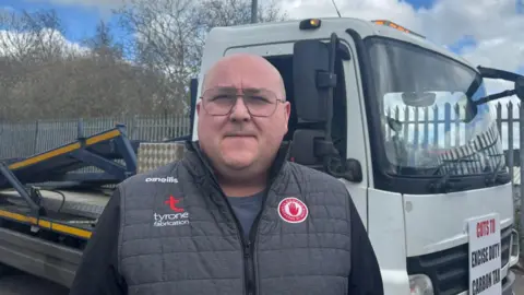 A bald man wearing glasses and a black long sleeve jacket with grey padding and a logo on the chest area stands in front of a recovery vehicle