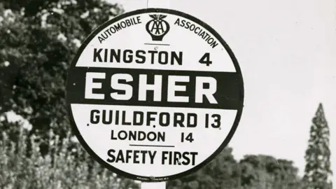 The AA A black-and-white image of a circular road sign for Esher, showing distances to Kingston, Guildford and London as well as the Automobile Association logo and the message "Safety First"