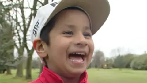 A five year old boy with a wide smile and a white baseball cap stands on a golf course, he is wearing a red top but only his head is pictured