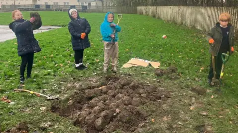 WCRT Four schoolchildren wearing outdoor clothing stand on green grass and mud. Some of the children have spades in their hands and others are holding up turf. 