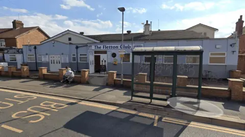 Google The Fletton Club is a one-storey building painted light blue with a bus stop in the foreground covered in graffiti.