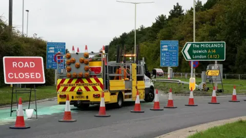 A Highways Mainenance vehicle can be seen alongside several cones and a sign saying 'Road closed'. Another sign points to the A14 and the Midlands. 